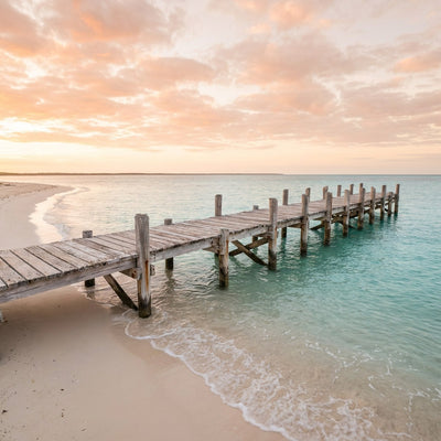 Beach house wall art, a wooden jetty reaching over calm turquoise water at sunrise beside pale sand