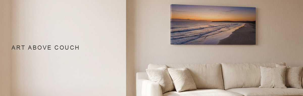 Art above couch: a living room with a long cream sofa, throw pillows and a wooden coffee table, with a wide beach-at-sunset canvas hung above the sofa